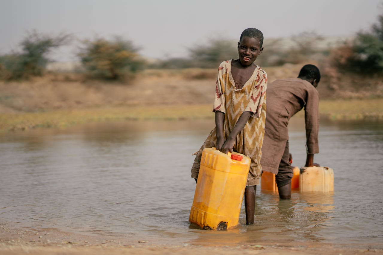 Two children gather water from a river using yellow containers, showcasing daily life.